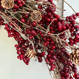 A close-up of a holiday wreath or decorative garland made of brown branches, glossy deep red berries, and natural tan pinecones against a plain light background.