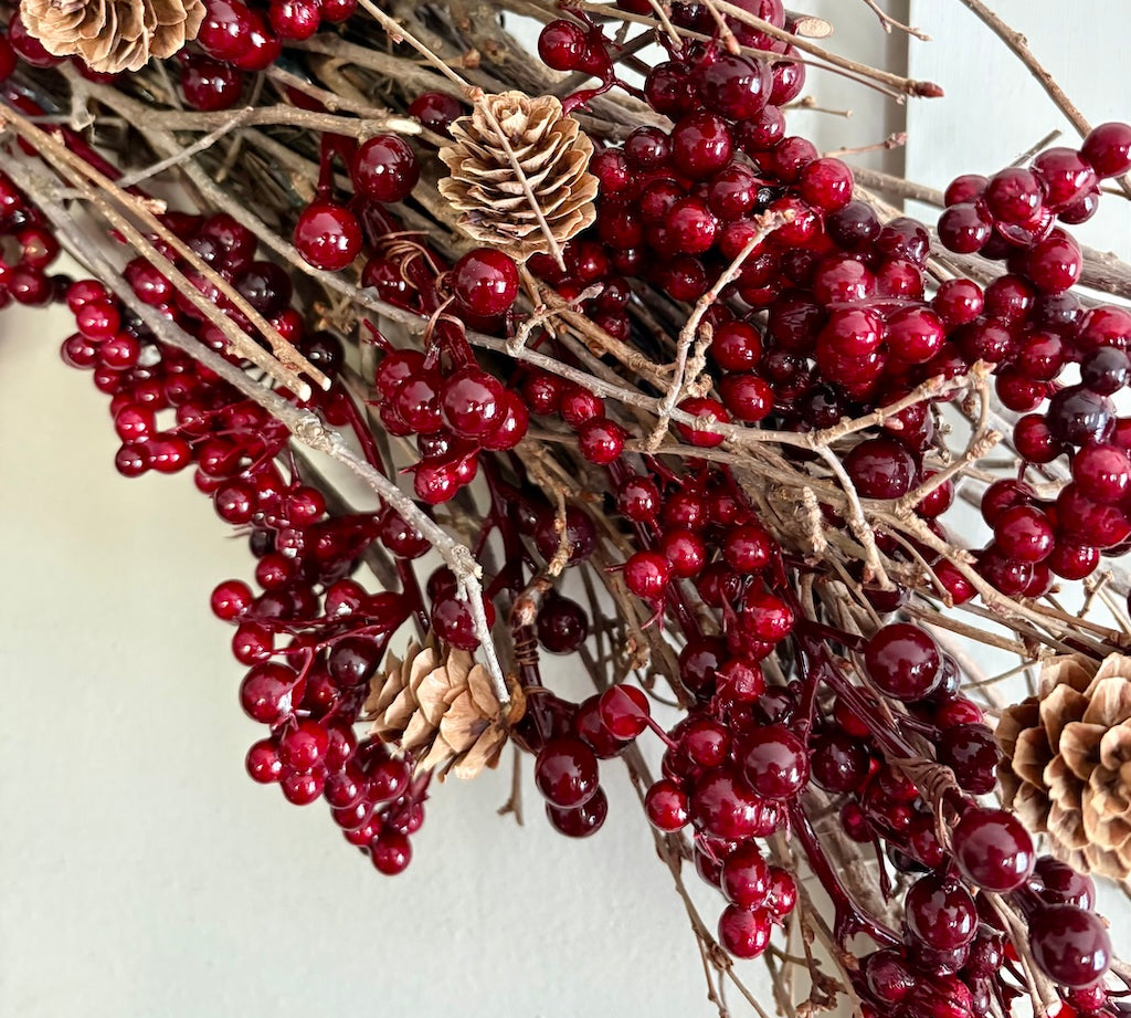 A close-up of a holiday wreath or decorative garland made of brown branches, glossy deep red berries, and natural tan pinecones against a plain light background.