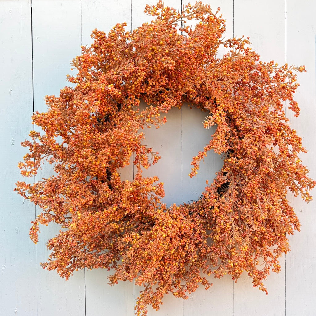 An autumn-themed artificial wreath with dense clusters of orange, rust, and yellow berries on a circular frame, displayed against a white painted wood plank background.