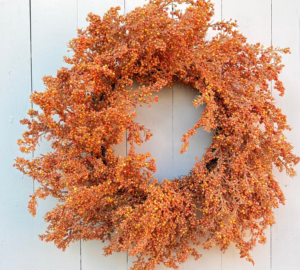 An autumn-themed artificial wreath with dense clusters of orange, rust, and yellow berries on a circular frame, displayed against a white painted wood plank background.
