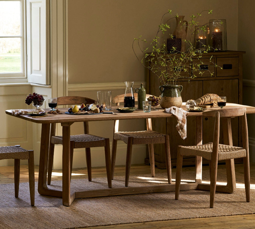A rustic wooden dining table with woven seat and back cushions, styled with glassware, bottles, and a vase of branches in a warm, sunlit room with beige walls and a window.