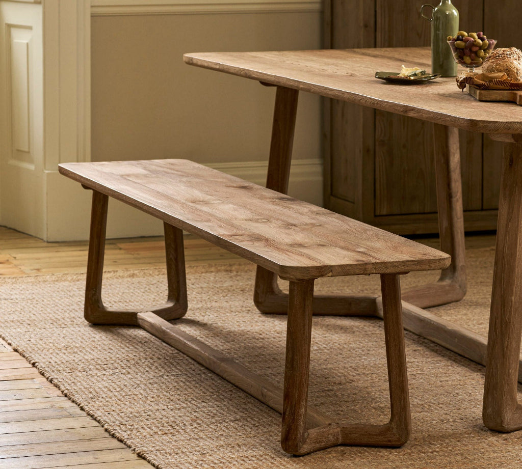 A rustic wooden dining table with matching bench set in a warm, neutral-toned dining room with a wood cabinet and neutral walls in the background.