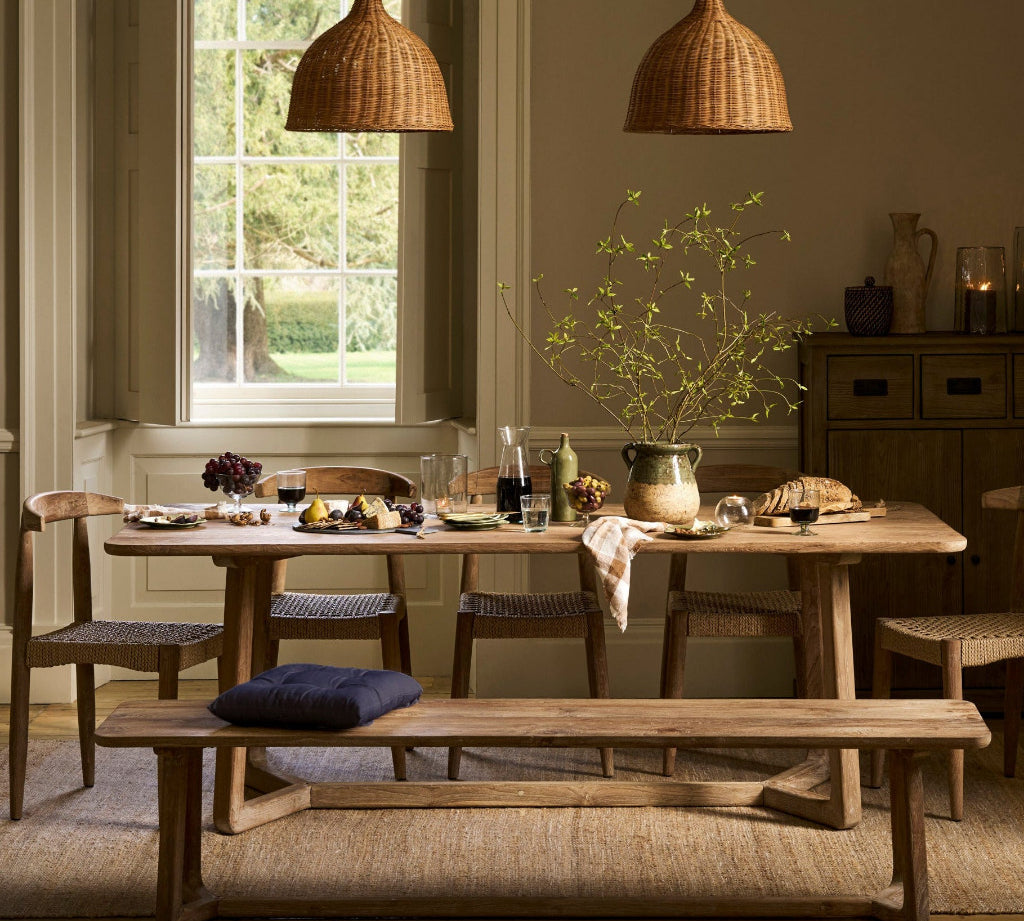 A rustic dining room scene with a wooden dining table, matching chairs and bench, two woven pendant lights, a large window with outdoor greenery, and a sideboard in the background.
