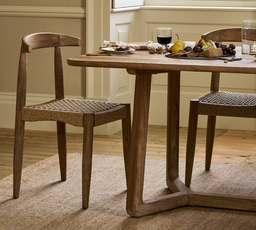 A rustic wooden dining table with two woven cane chairs in a bright, neutral-toned dining room setting.