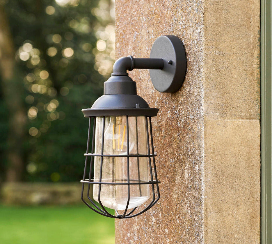 A dark metal outdoor wall sconce with a wire cage shade and visible warm bulb, mounted on a textured stone pillar with a blurred garden background.