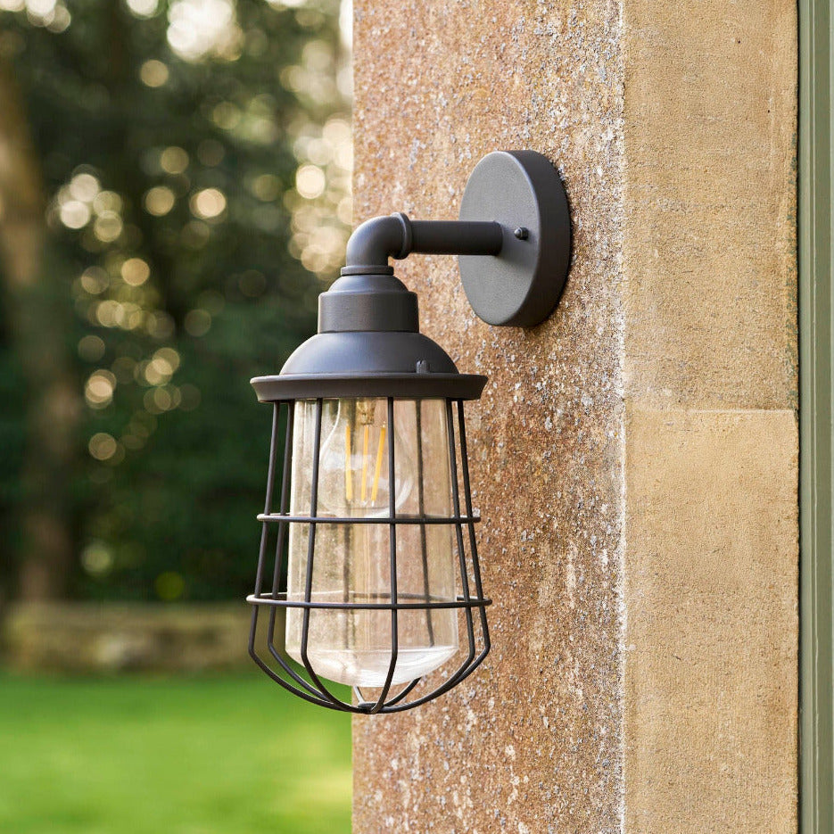 A dark metal outdoor wall sconce with a wire cage shade and visible warm bulb, mounted on a textured stone pillar with a blurred garden background.