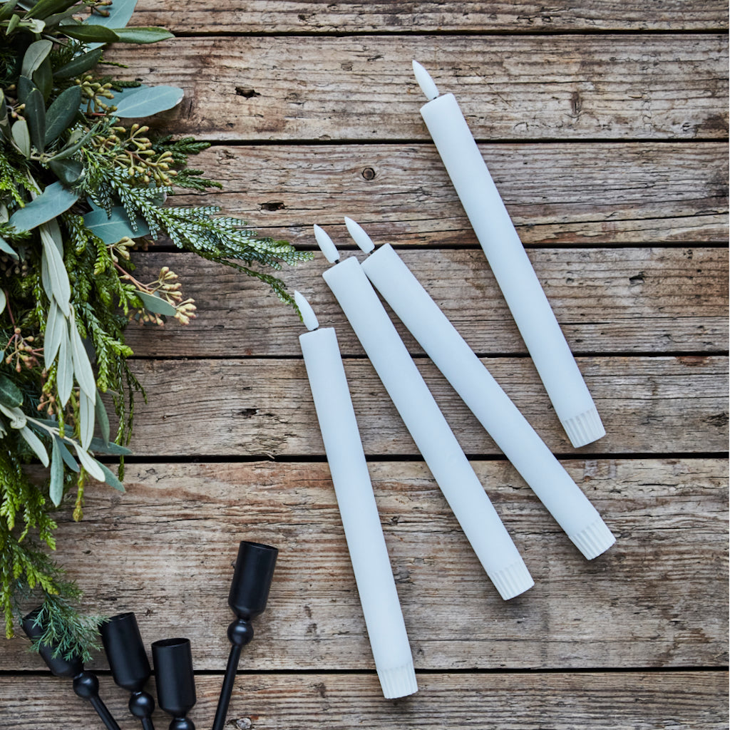 White candles and black candle holders on a wooden surface with greenery.