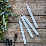 White candles and black candle holders on a wooden surface with greenery.