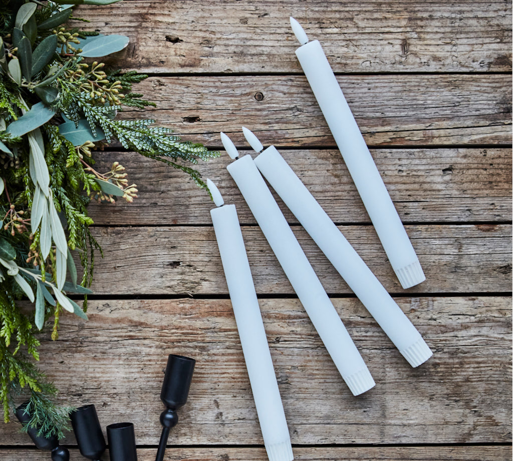 White candles and black candle holders on a wooden surface with greenery.