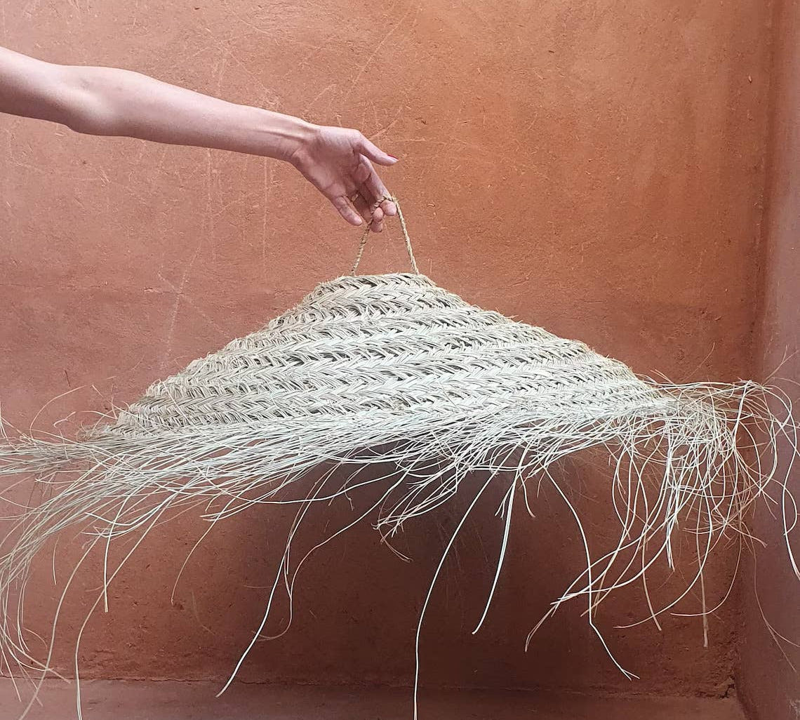 A hand holds up a large woven straw pendant light with long hanging frayed strands, set against a warm terracotta plaster wall and floor.