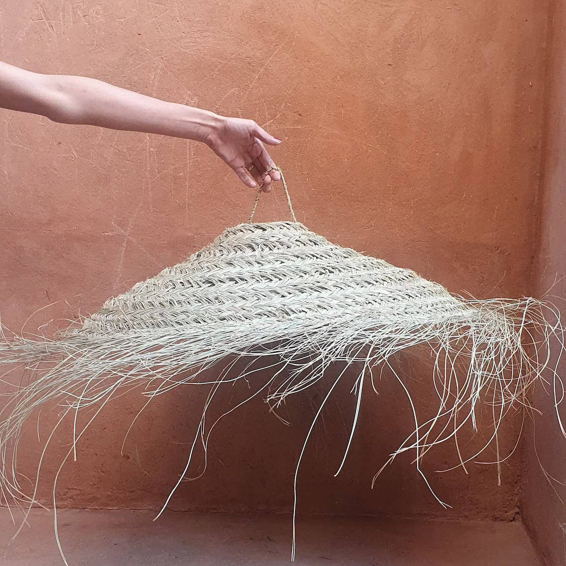 A hand holds up a large woven straw pendant light with long hanging frayed strands, set against a warm terracotta plaster wall and floor.