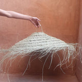 A hand holds up a large woven straw pendant light with long hanging frayed strands, set against a warm terracotta plaster wall and floor.
