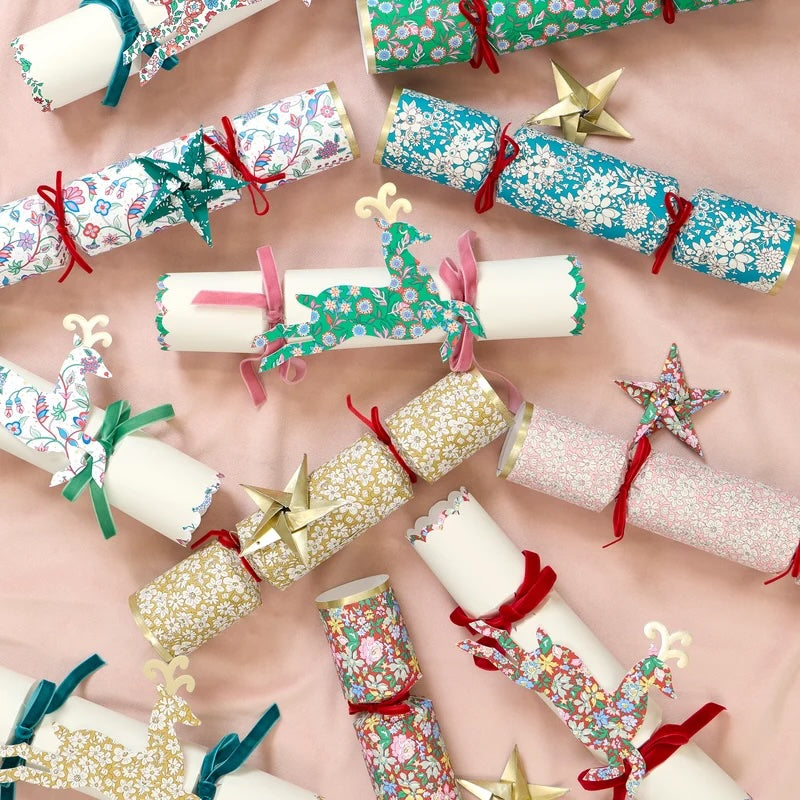 A styled flat-lay of Christmas cracker packs wrapped in festive patterned paper, tied with red, pink, and green ribbons, and decorated with gold reindeer and star ornaments on a soft pink background.