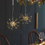 Two spherical starburst LED pendant lights with thin metallic wire spikes glow warmly from above in a dining room setting with dark paneled walls and wooden chairs.