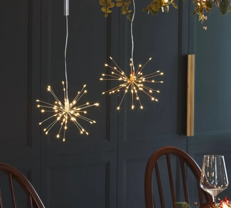 Two spherical starburst LED pendant lights with thin metallic wire spikes glow warmly from above in a dining room setting with dark paneled walls and wooden chairs.