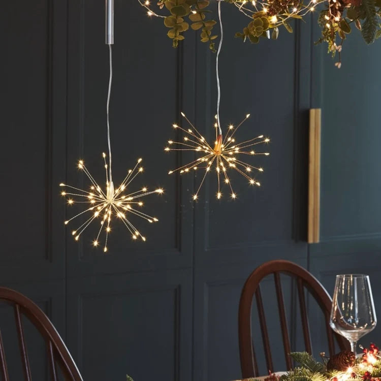 Two spherical starburst LED pendant lights with thin metallic wire spikes glow warmly from above in a dining room setting with dark paneled walls and wooden chairs.