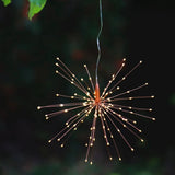 A hanging starburst-style LED light with many thin wire arms and small glowing bulbs, suspended against a dark outdoor background with blurred greenery.