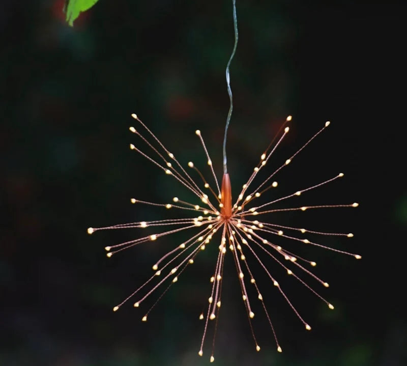 A hanging starburst-style LED light with many thin wire arms and small glowing bulbs, suspended against a dark outdoor background with blurred greenery.