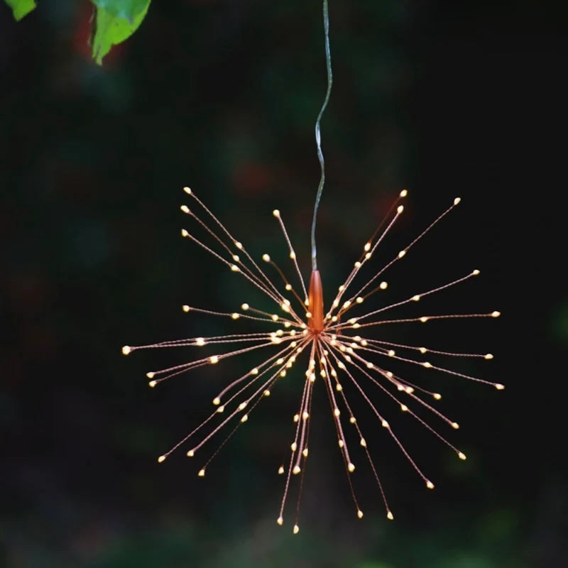 A hanging starburst-style LED light with many thin wire arms and small glowing bulbs, suspended against a dark outdoor background with blurred greenery.