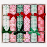 A boxed set of six Christmas crackers with festive patterned paper rolls tied with red and green ribbon bows, arranged in a light pink gift box on a white background.