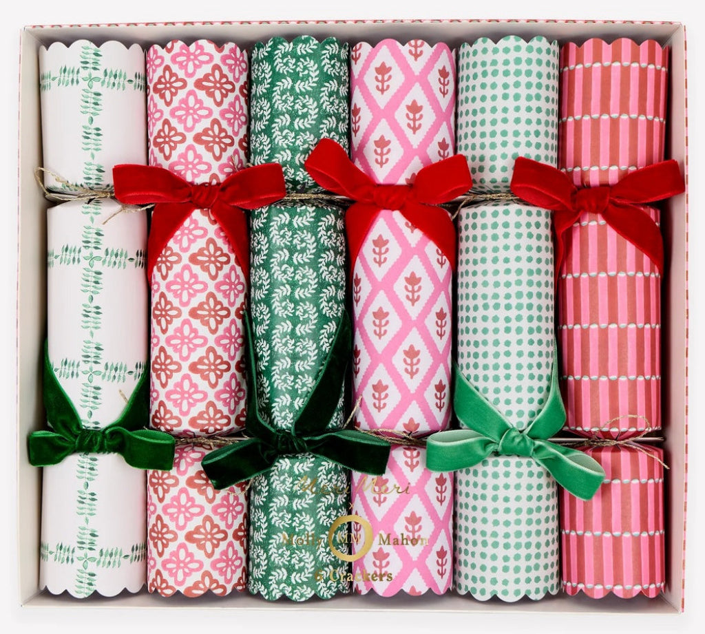 A boxed set of six Christmas crackers with festive patterned paper rolls tied with red and green ribbon bows, arranged in a light pink gift box on a white background.