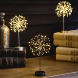 Lightstyle dandelion table lights in all three colourways — silver, black and gold — glowing with warm white LEDs on a wooden surface with books and a mercury glass bottle