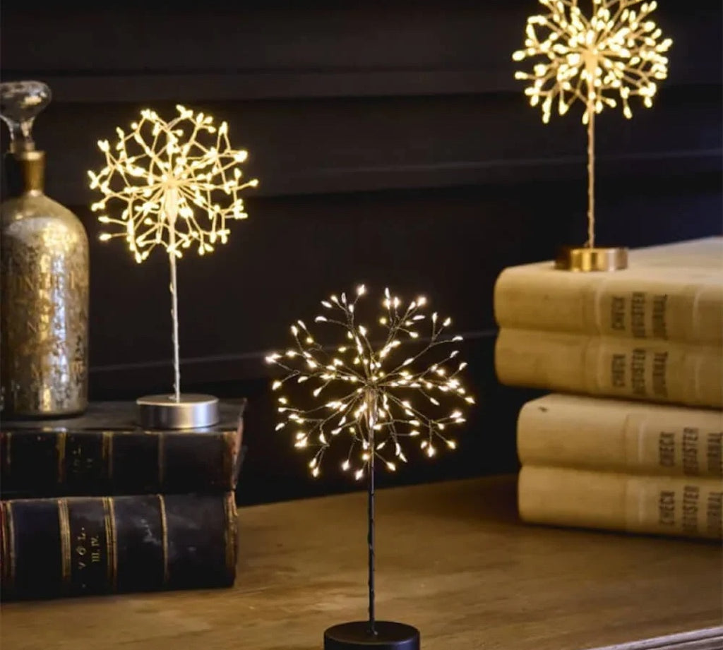 Lightstyle dandelion table lights in all three colourways — silver, black and gold — glowing with warm white LEDs on a wooden surface with books and a mercury glass bottle