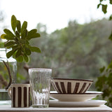 Dining table setting with striped mug, glass, and bowl against a blurred green outdoor background