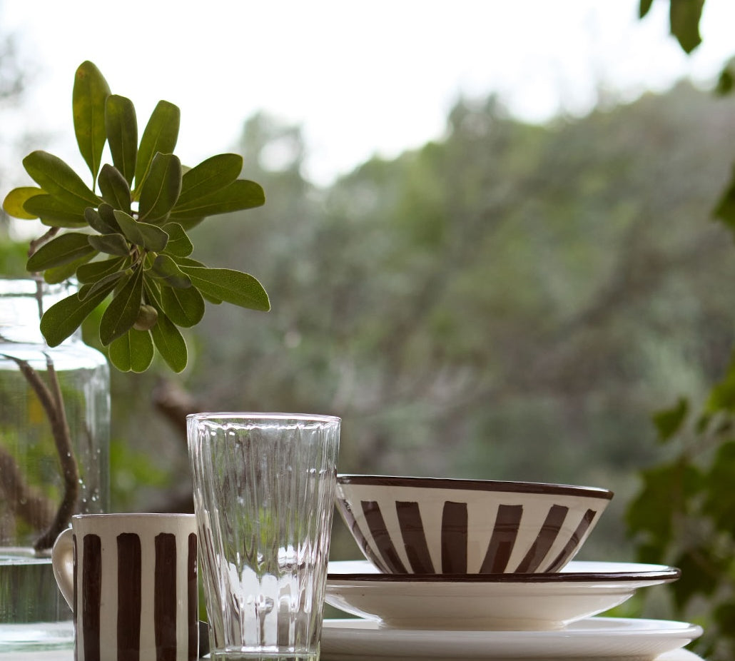 Dining table setting with striped mug, glass, and bowl against a blurred green outdoor background