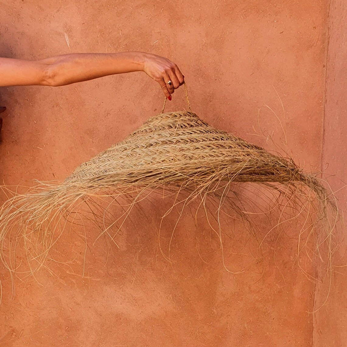 A hand holds the top of a large woven straw lampshade or decorative hanging against a warm terracotta stucco wall.