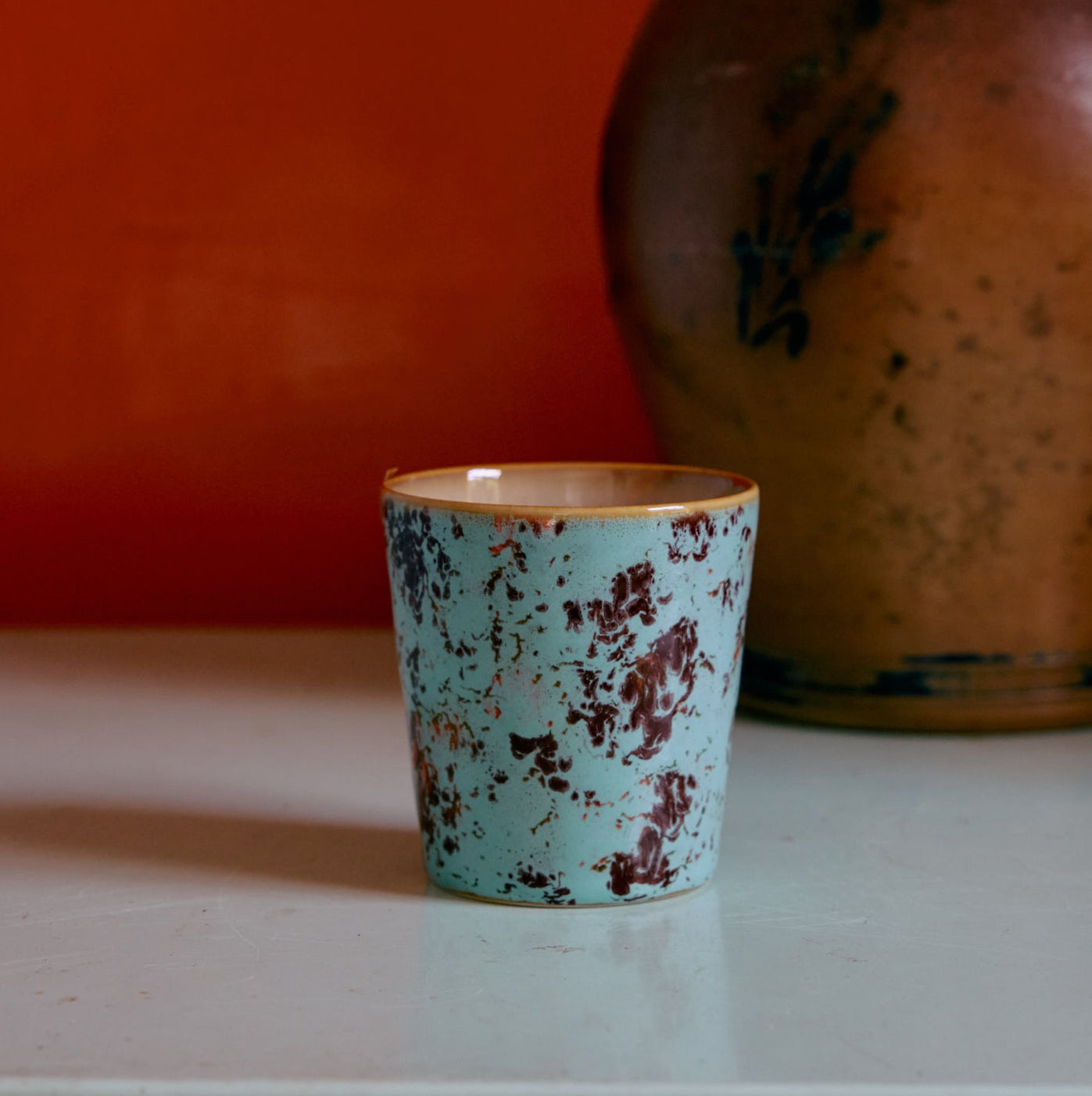 Teal ceramic cup with brown interior on a white surface against an orange wall