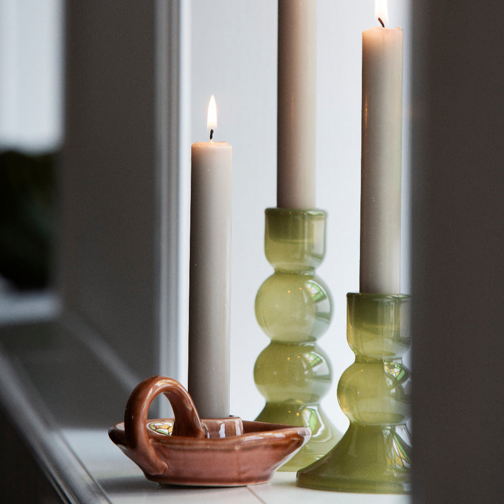 Three lit taper candles displayed on a windowsill, including two in translucent olive-green glass holders and one in a small terracotta-colored ceramic boat-shaped holder.