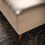Close-up of a rectangular upholstered ottoman or bench corner, showing a light beige textured fabric seat and back with a small wooden footed leg, placed on a dark patterned rug.