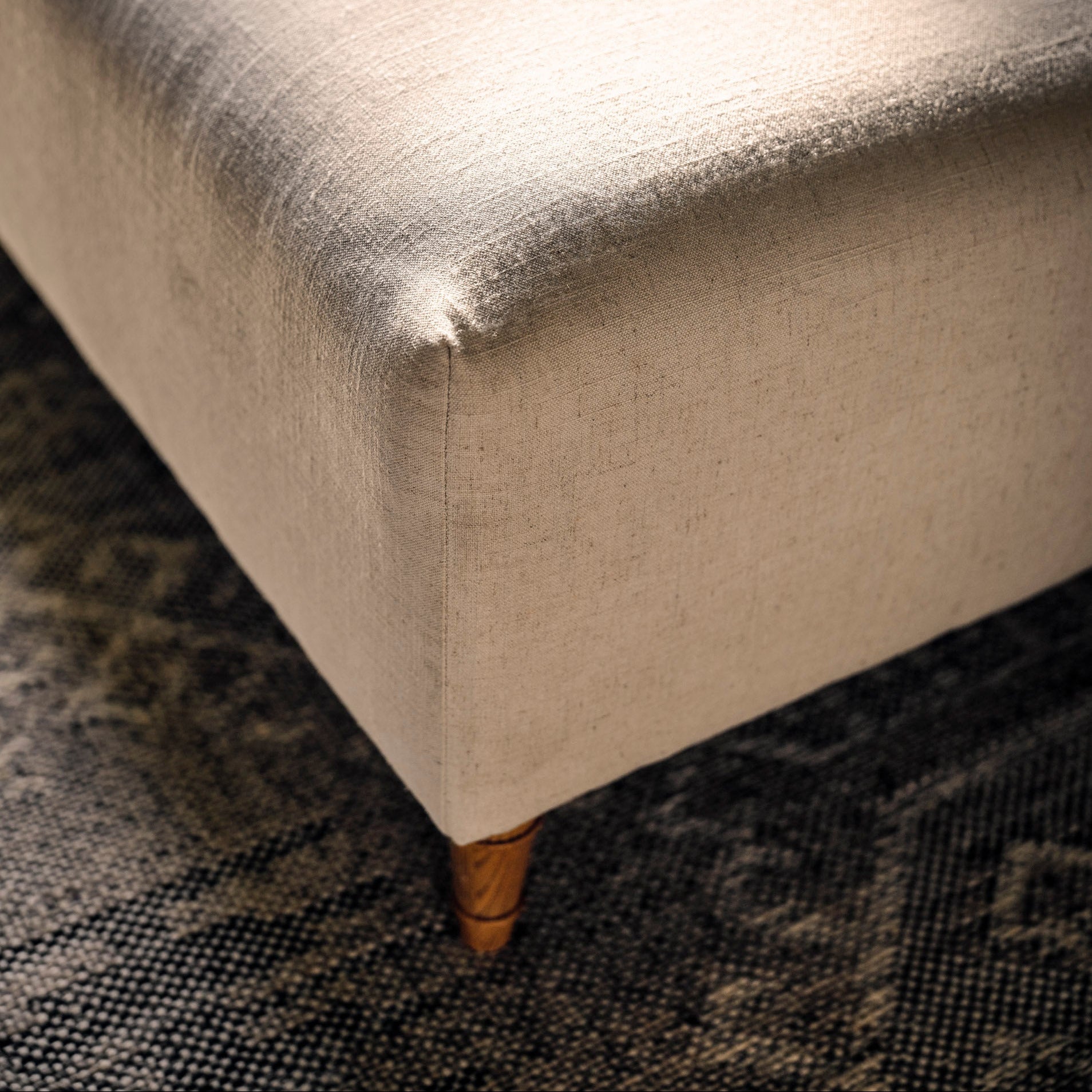 Close-up of a rectangular upholstered ottoman or bench corner, showing a light beige textured fabric seat and back with a small wooden footed leg, placed on a dark patterned rug.