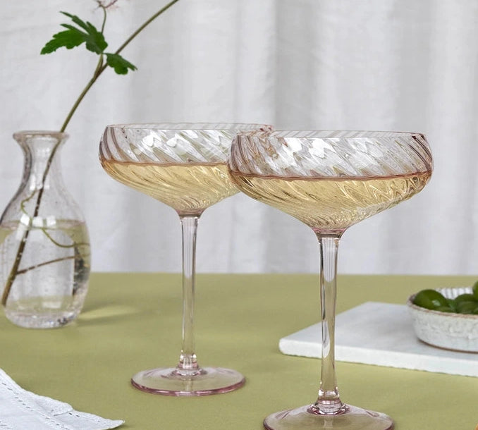 Two pink-tinted coupe glasses with a textured, ribbed pattern, filled with white wine, set on a green tablecloth with a small glass vase and olives in the background.