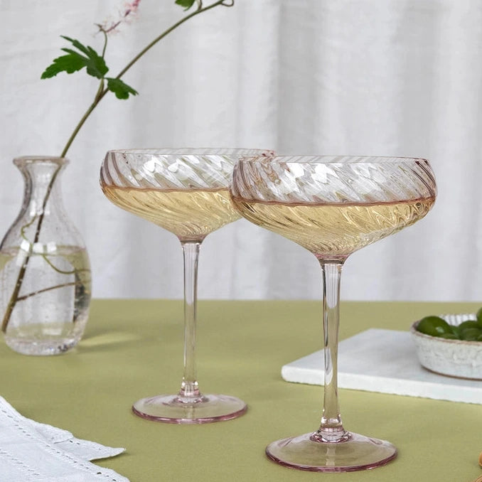 Two pink-tinted coupe glasses with a textured, ribbed pattern, filled with white wine, set on a green tablecloth with a small glass vase and olives in the background.