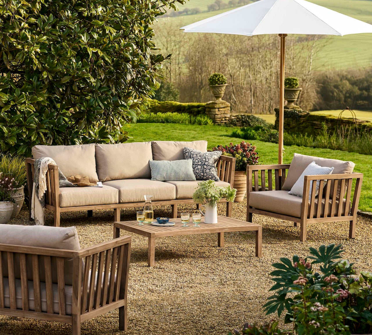 Outdoor patio set with beige cushions, wooden furniture, and a white umbrella in a garden setting.