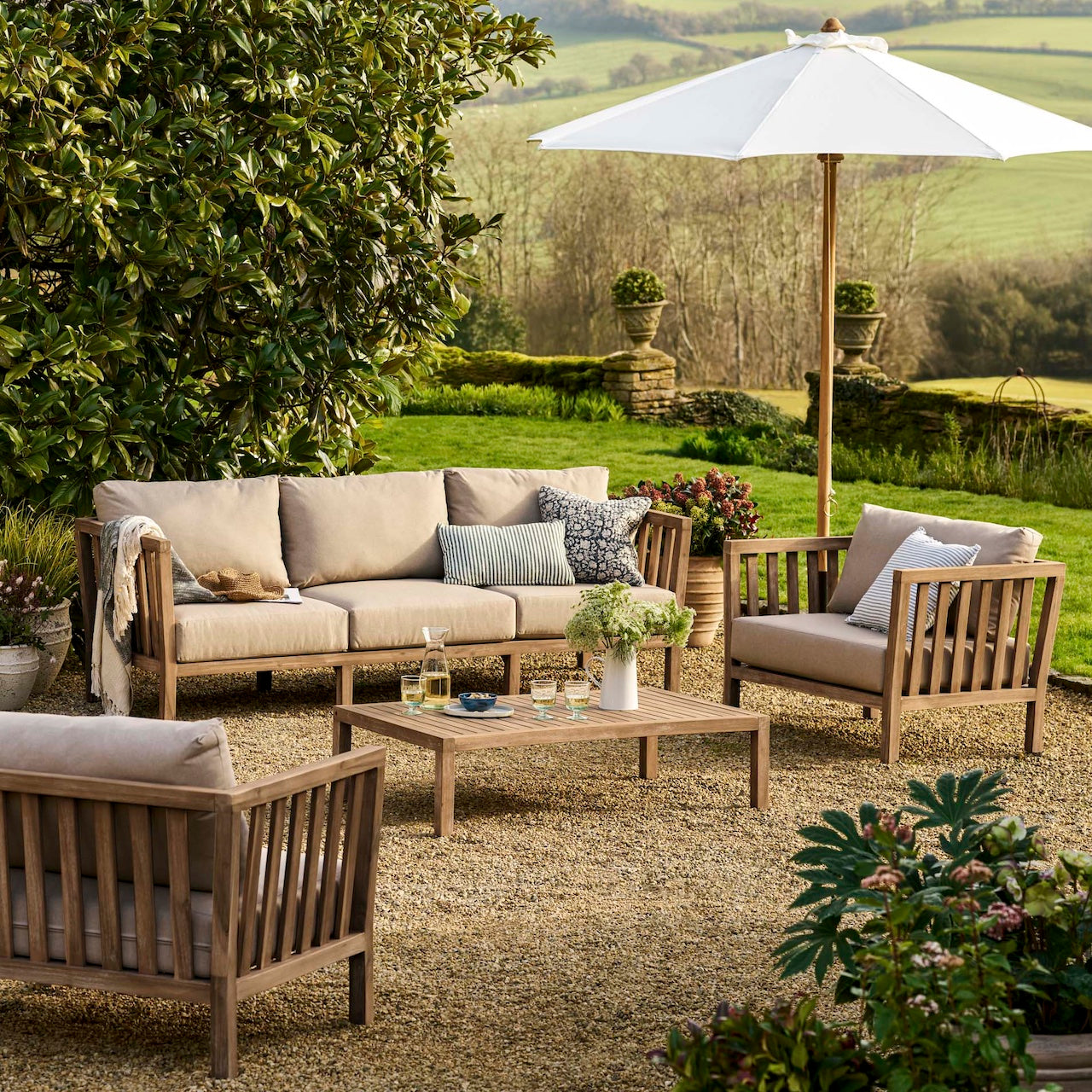 Outdoor patio set with beige cushions, wooden furniture, and a white umbrella in a garden setting.