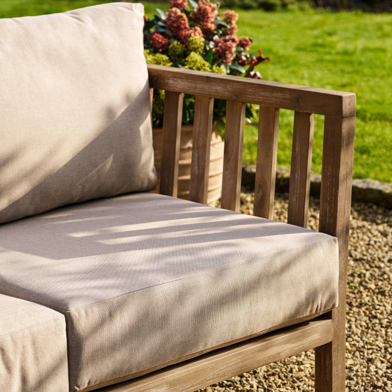 Wooden outdoor sofa with beige cushions on a pebble path with greenery in the background