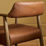 Close-up of a modern wooden dining chair with warm brown leather upholstery on the seat and backrest, set against a neutral beige wall and wood floor.