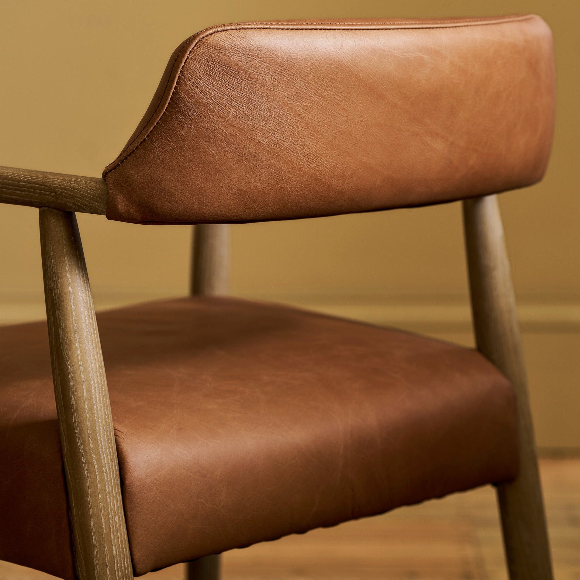 Close-up of a modern wooden dining chair with warm brown leather upholstery on the seat and backrest, set against a neutral beige wall and wood floor.