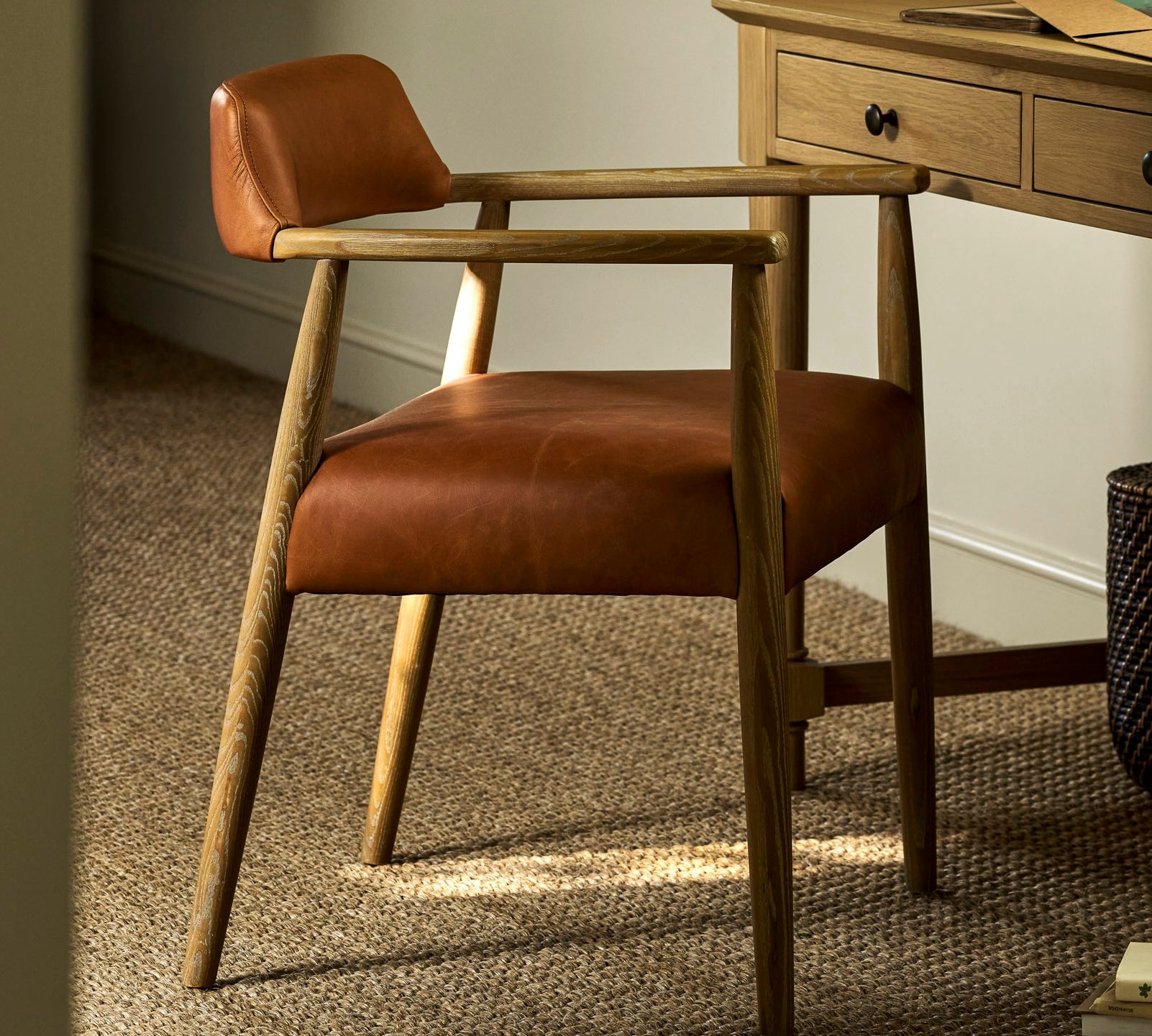 A mid-century modern wooden armchair with warm brown leather upholstery, positioned in a home office setting next to a wooden desk on a textured beige rug.