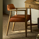 A mid-century modern wooden armchair with warm brown leather upholstery, positioned in a home office setting next to a wooden desk on a textured beige rug.