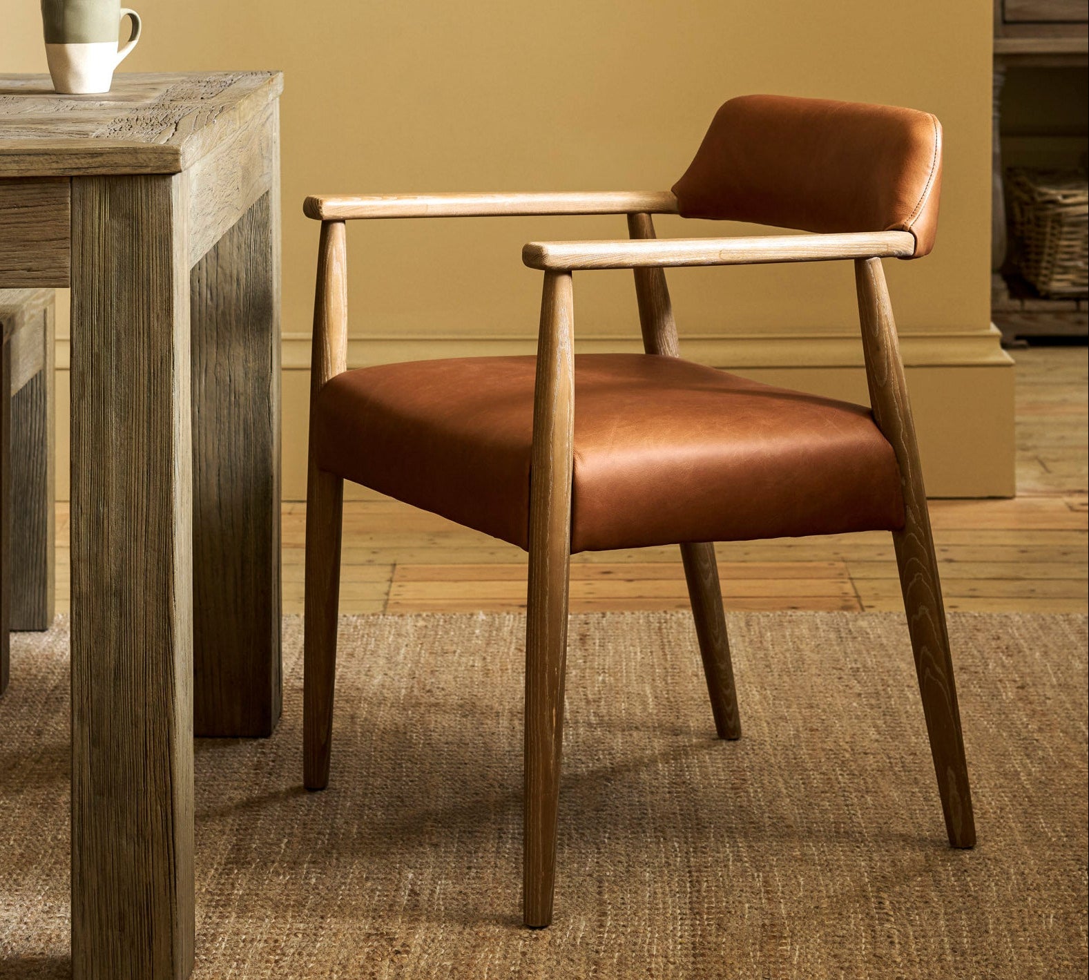A mid-century modern accent chair with a warm brown leather seat and backrest, supported by a light natural wood frame, positioned next to a rustic wooden dining table in a warm, neutral-toned room.