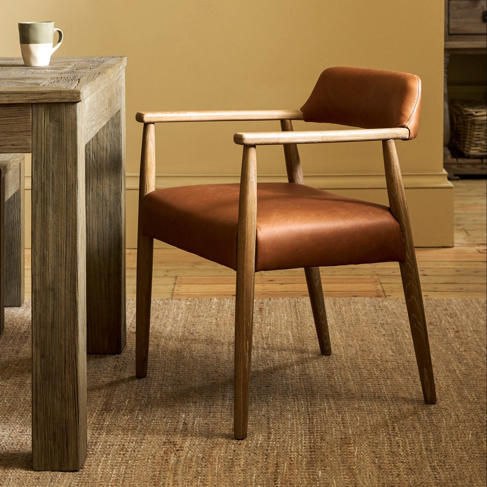 A mid-century modern accent chair with a warm brown leather seat and backrest, supported by a light natural wood frame, positioned next to a rustic wooden dining table in a warm, neutral-toned room.