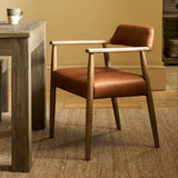 A mid-century modern accent chair with a warm brown leather seat and backrest, supported by a light natural wood frame, positioned next to a rustic wooden dining table in a warm, neutral-toned room.