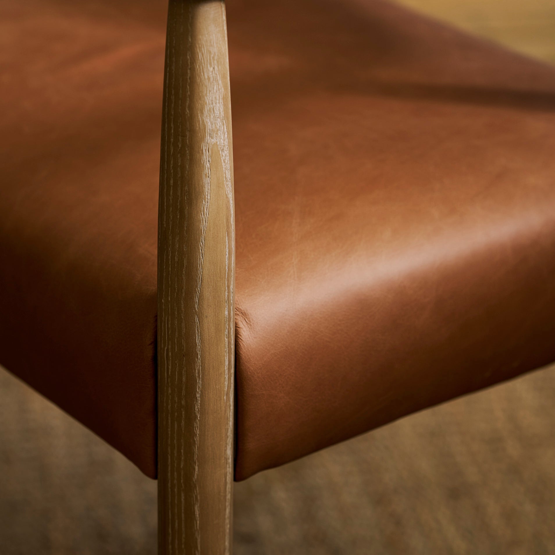 Close-up of a wooden armchair with a warm brown leather seat cushion, showing the armrest and leg details against a softly blurred neutral background.
