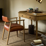 A mid-century modern writing desk and matching armchair in a warm wood and brown leather set, styled in a cozy, sunlit attic study with a brass desk lamp and books.