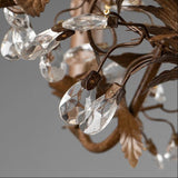 Close-up of a decorative chandelier or light fixture with faceted clear crystal teardrop pendants and brown metal leaf-shaped arms on a gray background.