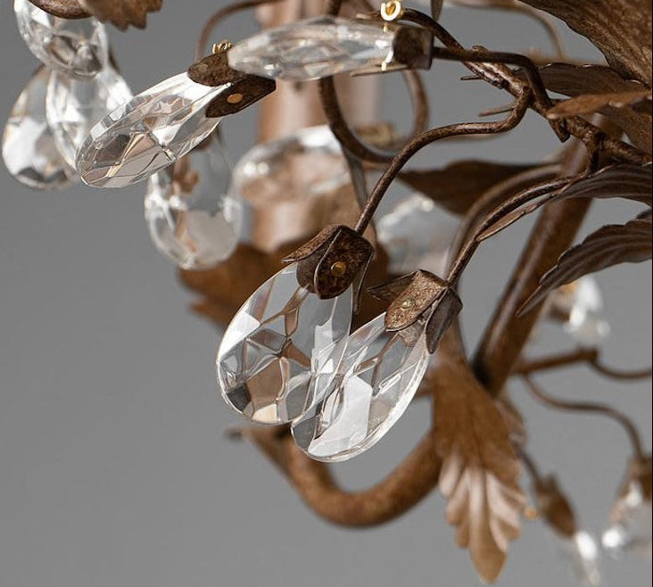Close-up of a decorative chandelier or light fixture with faceted clear crystal teardrop pendants and brown metal leaf-shaped arms on a gray background.